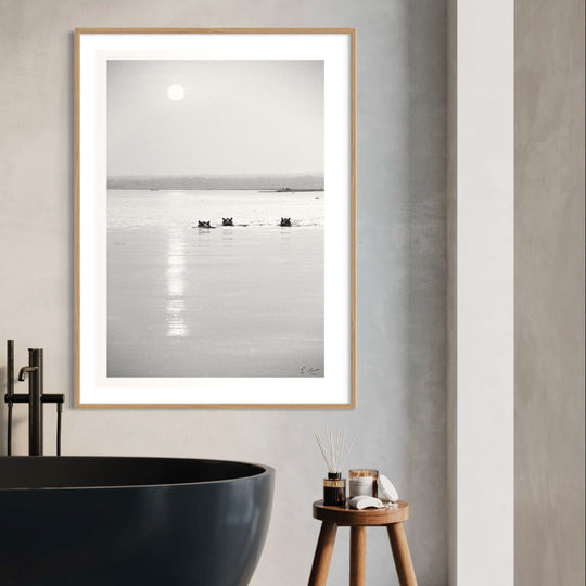 A black and white photograph of hippos in a lagoon at sunset, displayed as a wall art print above a wooden stool in a room with a bathtub and candles.